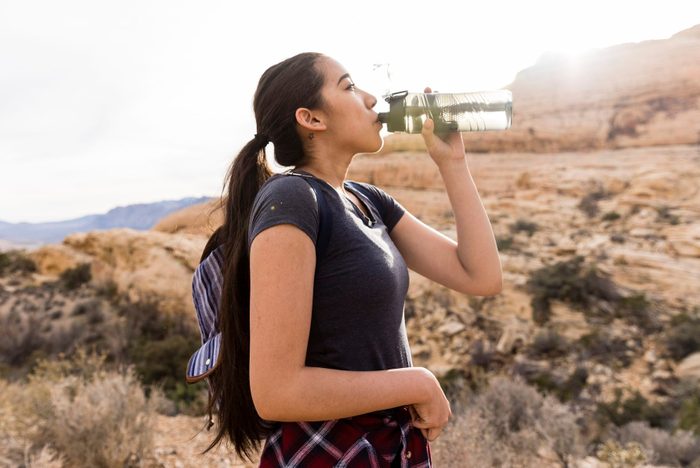Woman with backpack drinking water while standing by rock formation against clear sky