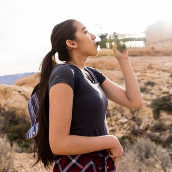 Woman with backpack drinking water while standing by rock formation against clear sky