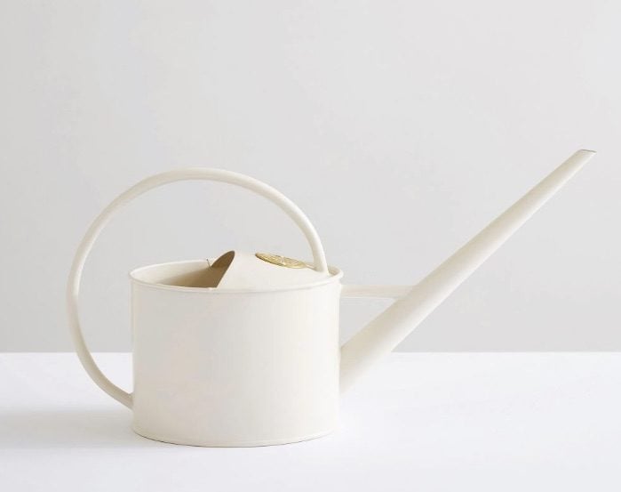 A matte white watering can with long neck, circular handle and gold emblem on top.