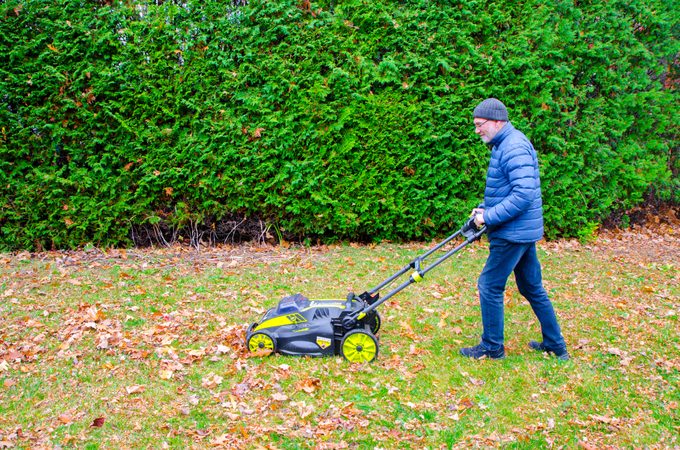 Man passing lawn mower over autumn leaves on grass